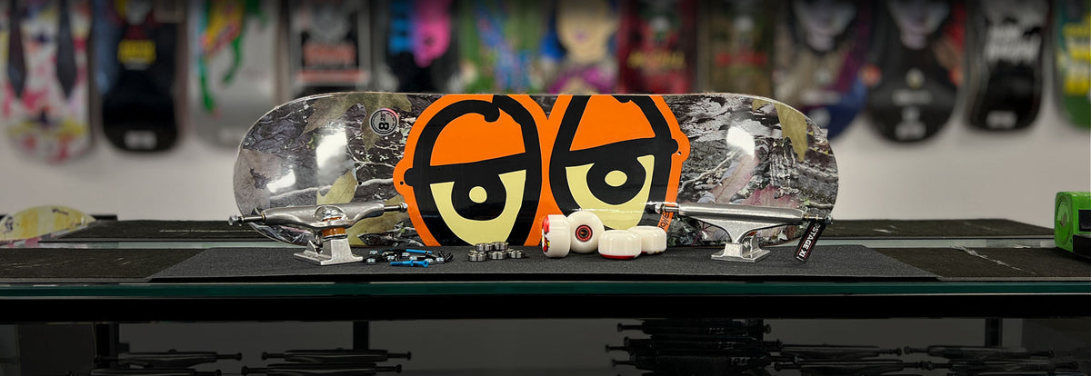 A complete skateboard deck with a clear underside and a bold orange and black cartoon face graphic, displayed on a black table with other skateboard parts and blurred skateboards in the background.